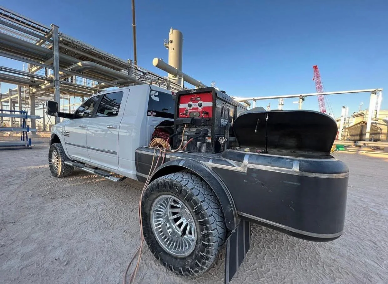 White pickup converted into a mobile welding rig with a welder, leads, storage compartments, and industrial piping behind it.