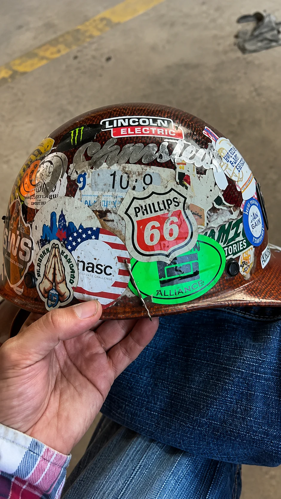 Hand holding a worn hard hat covered in jobsite and trade stickers.
