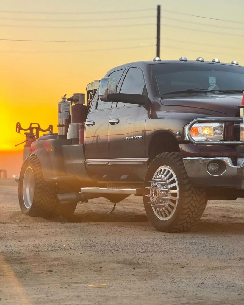 Black heavy-duty pickup truck with large wheels photographed on a dirt lot at sunset.